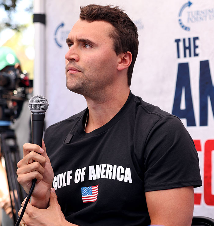 Man holding microphone wearing black Gulf of America shirt discussing Charlie Kirk life homework assignment controversy