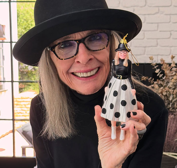 Diane Keaton smiling, wearing a black hat and glasses, holding a doll dressed in a black and white polka dot outfit.