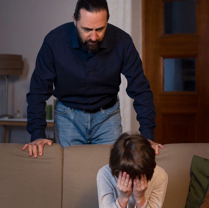 Man displaying cruel behavior towards a distressed child covering his face on a couch, revealing cruelty and emotional harm.