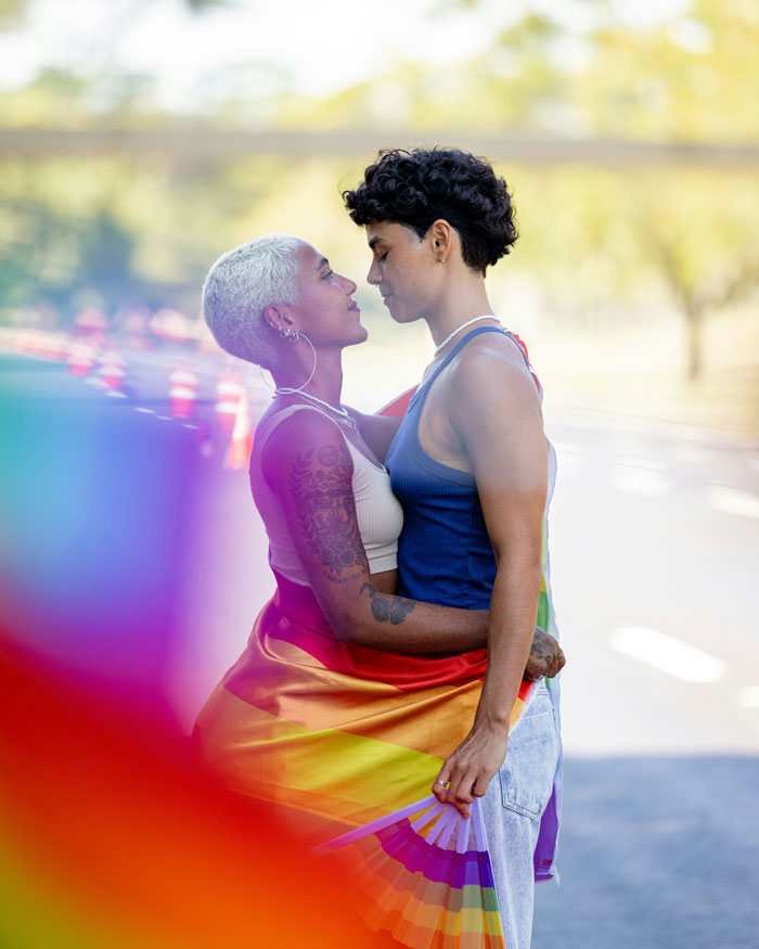 Two women embracing outdoors with a rainbow flag, highlighting lesbian perspectives amid men-only gay bar controversy.