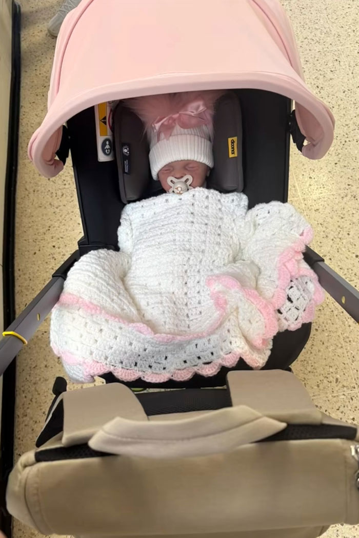 Baby doll in a car seat covered by a white blanket with a pink trim, highlighting woman's outrageous 10-month lie discovery.