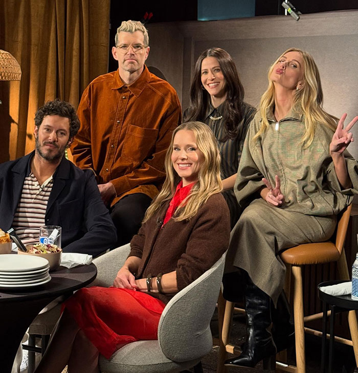Group of five people sitting and posing indoors, featuring Kristen Bell in a red dress amid backlash discussions.
