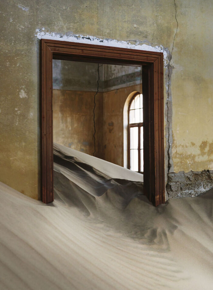 Desert sand filling an abandoned home through a doorway with cracked walls and a tall window in the background.