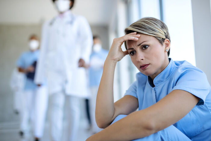 Nurse sitting on hospital floor looking stressed with blurred medical staff in background, capturing hospital work atmosphere.