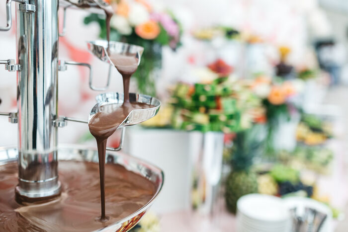 Chocolate fountain flowing with smooth chocolate, surrounded by blurred fruit and dessert arrangements in a bright setting.