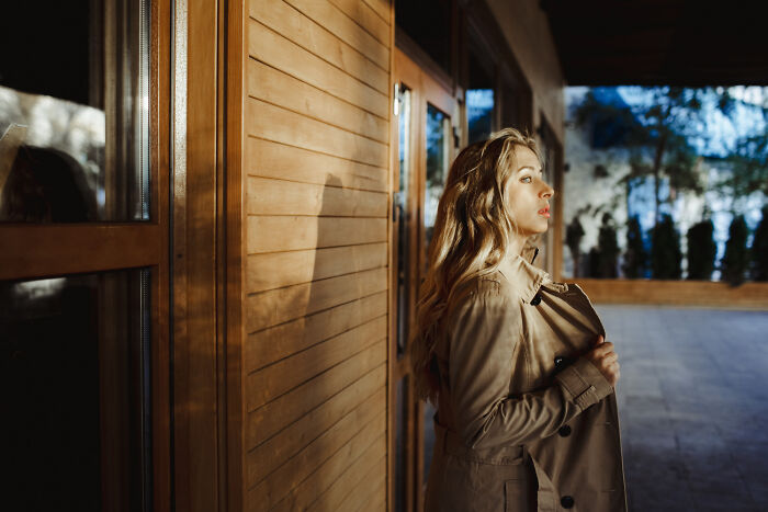 Woman in a beige trench coat standing outside a wooden building at dusk, evoking themes of stalking and observation.