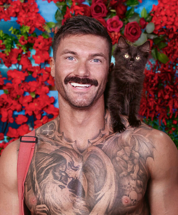 Shirtless Australian firefighter with tattoos smiles holding a black kitten on his shoulder against red floral background