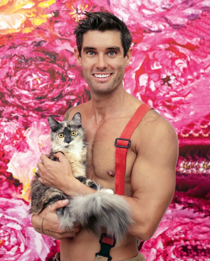 Shirtless Australian firefighter wearing suspenders holding a fluffy kitten against a pink floral background for calendar shoot.