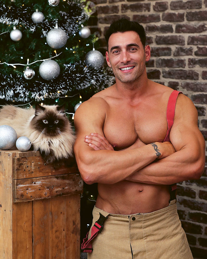 Muscular Australian firefighter posing shirtless next to a fluffy cat by a decorated Christmas tree for firefighters calendar.