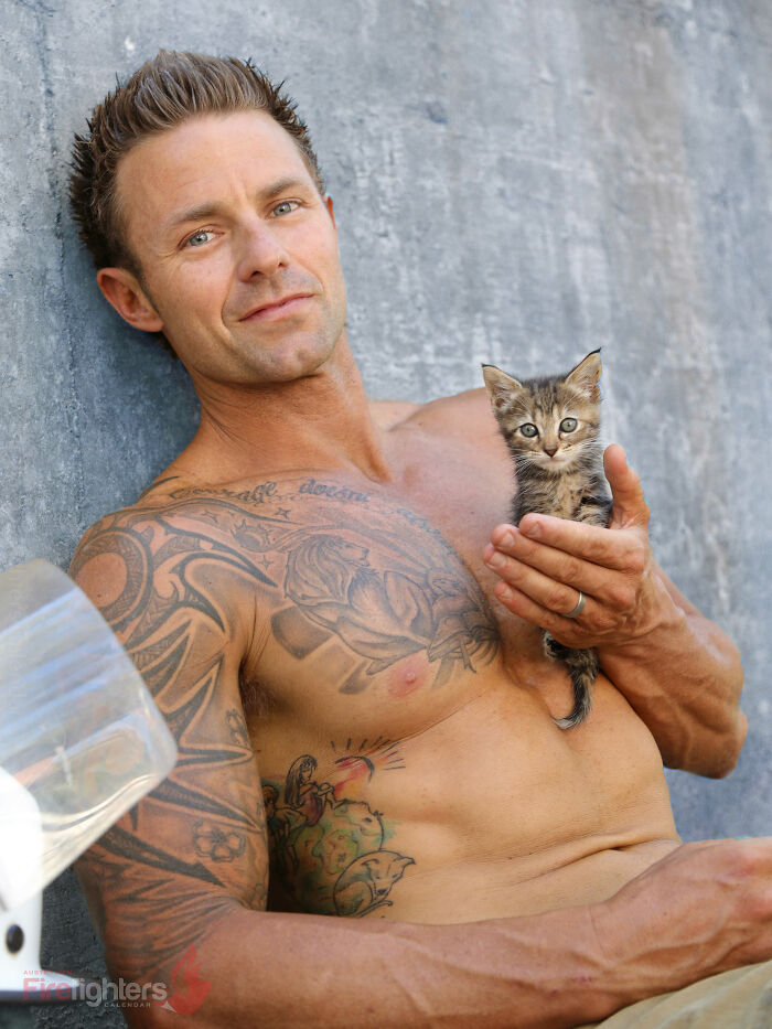 Shirtless Australian firefighter with tattoos holding a small kitten, featured in a firefighters calendar photo shoot.