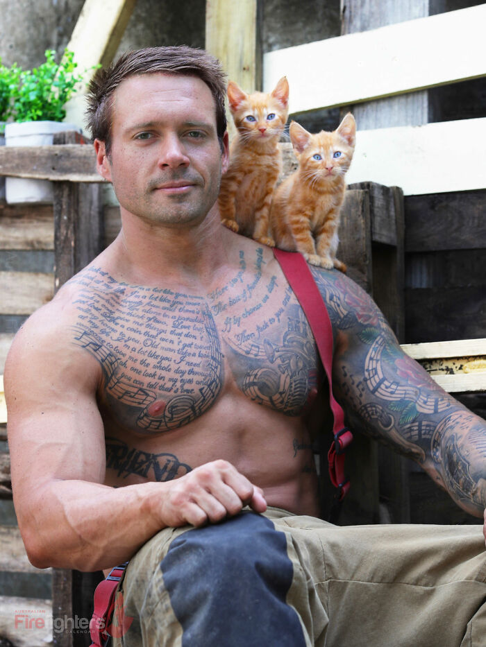 Australian firefighter with tattoos sitting outdoors with two orange kittens on his shoulder for the firefighters calendar.