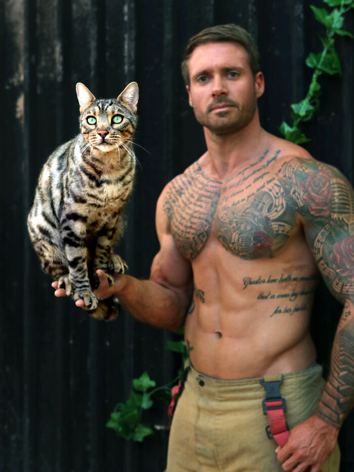 Shirtless tattooed Australian firefighter holding a spotted kitten in a calendar photoshoot setting.