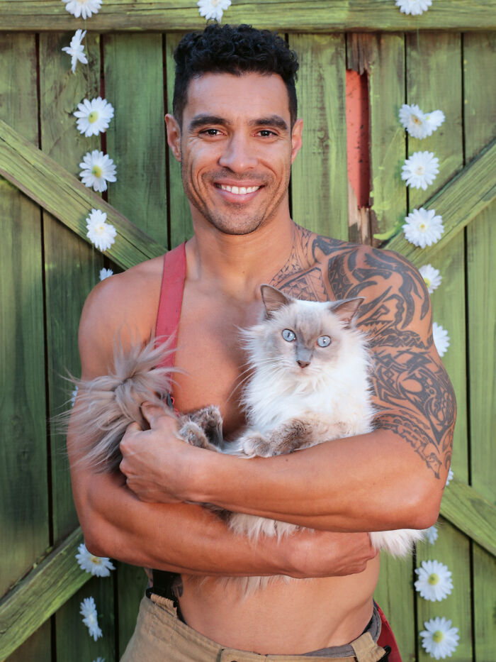 Smiling firefighter with tattooed arm holding a fluffy kitten in front of a green wooden fence with white flowers.