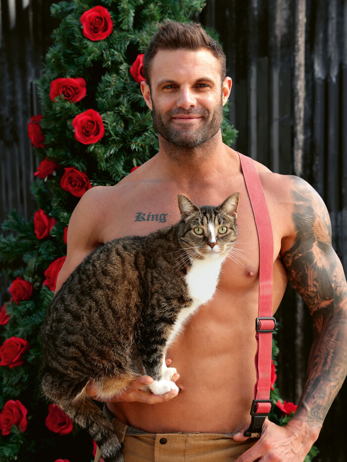 Shirtless firefighter with a tattoo holds a tabby cat in front of a rose-decorated tree for Australian firefighters calendar.