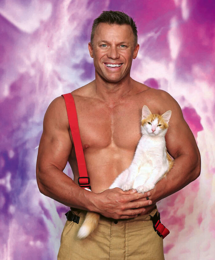 Shirtless Australian firefighter holding an orange and white kitten against a purple-pink background for calendar photoshoot.