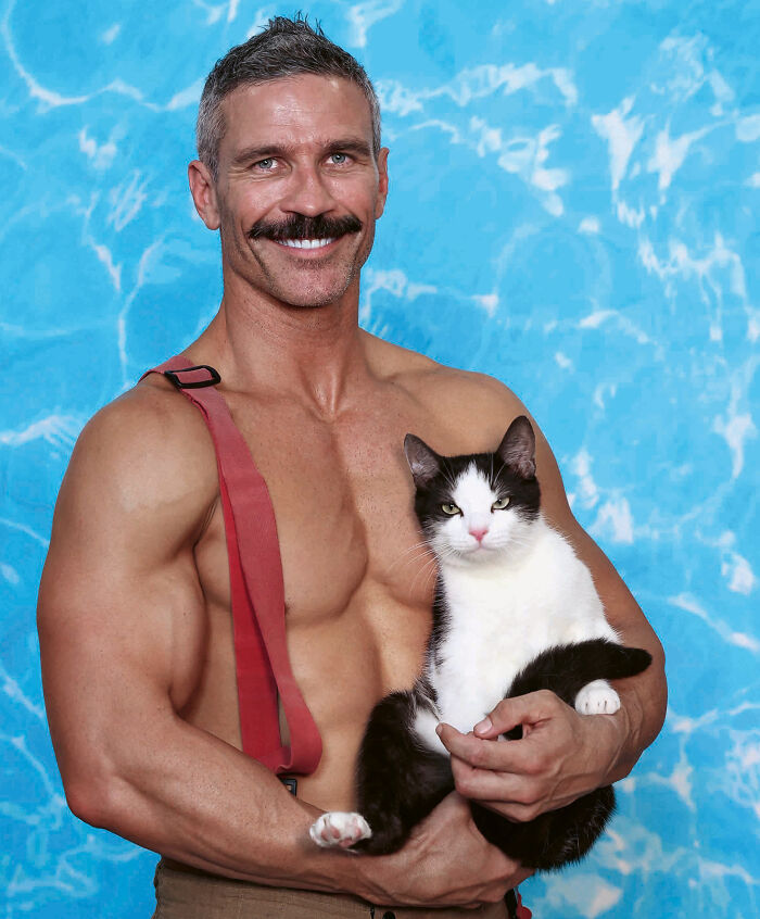 Shirtless firefighter from Australian Firefighters Calendar holding a black and white kitten against blue backdrop.