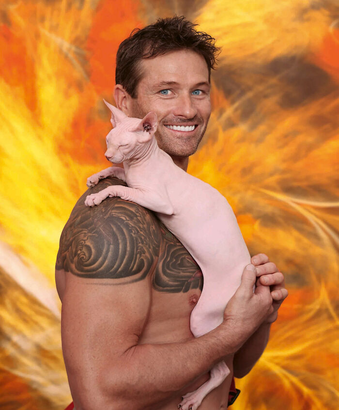 Shirtless Australian firefighter with tattoos holding a hairless kitten against a fiery orange background for calendar shoot