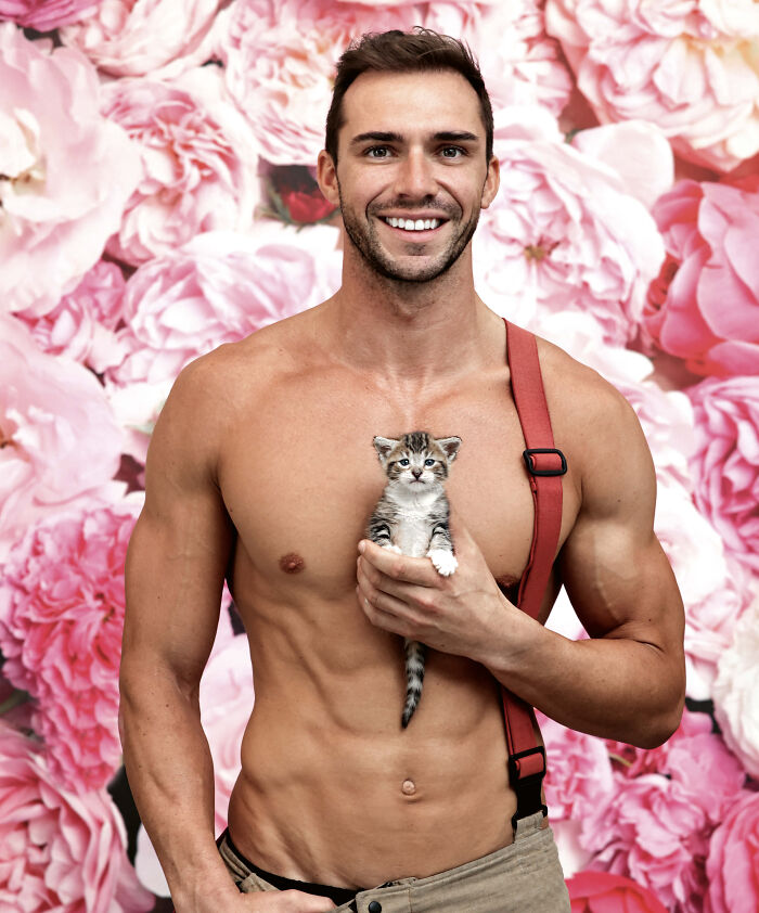 Shirtless Australian firefighter holding a kitten against a pink floral background for a firefighter calendar photoshoot.
