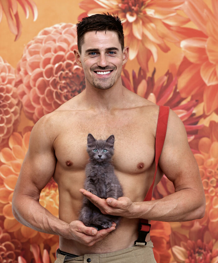 Shirtless Australian firefighter smiling and holding a gray kitten against a vibrant floral background for a calendar.