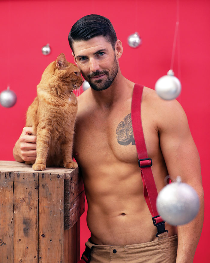 Shirtless Australian firefighter with suspenders posing with an orange kitten on a wooden crate against a red background.