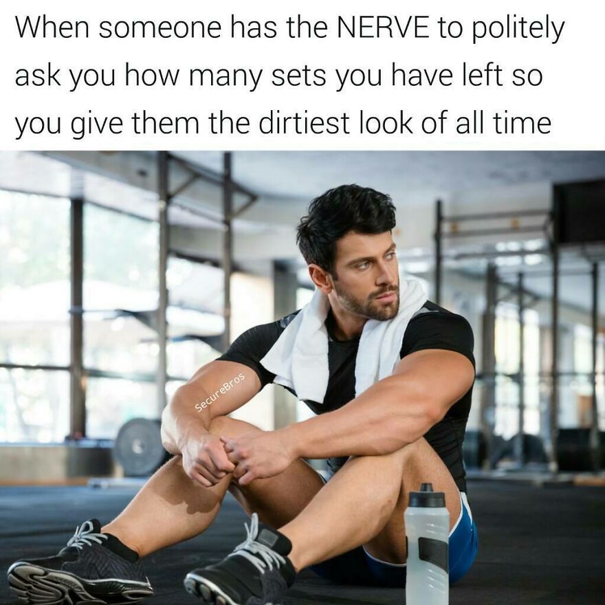 Muscular man sitting on gym floor with towel around neck, giving a dirty look after being asked about his workout sets.