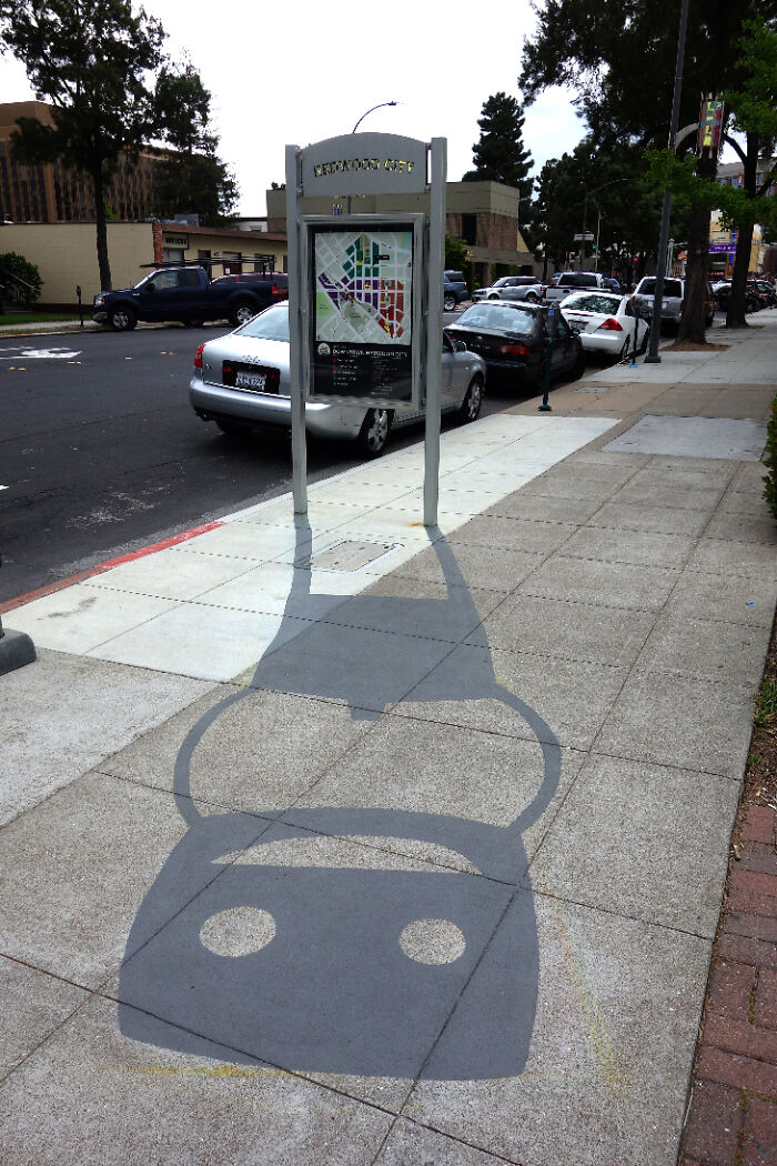 Shadow art installation on sidewalk transforming a street sign's shadow into a playful robot face.
