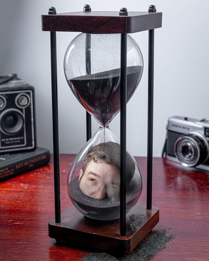Surreal photo manipulation showing a man's face trapped inside an hourglass filled with black sand on a wooden table.