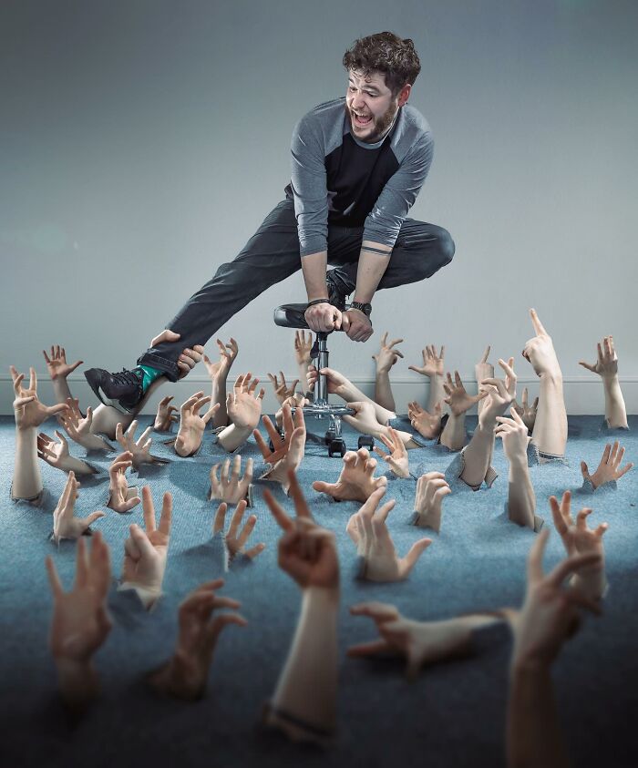Man balancing on a chair above numerous hands emerging from the floor in an amazing logic-challenging photo manipulation.