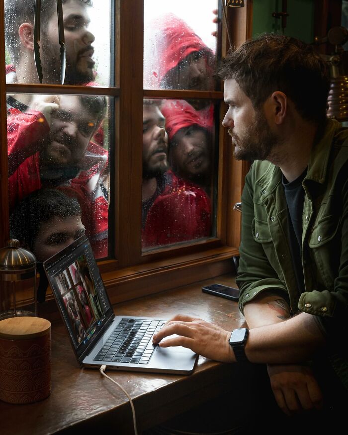 Man creating amazing logic-challenging photo manipulations on laptop while multiple faces appear outside window on rainy day.