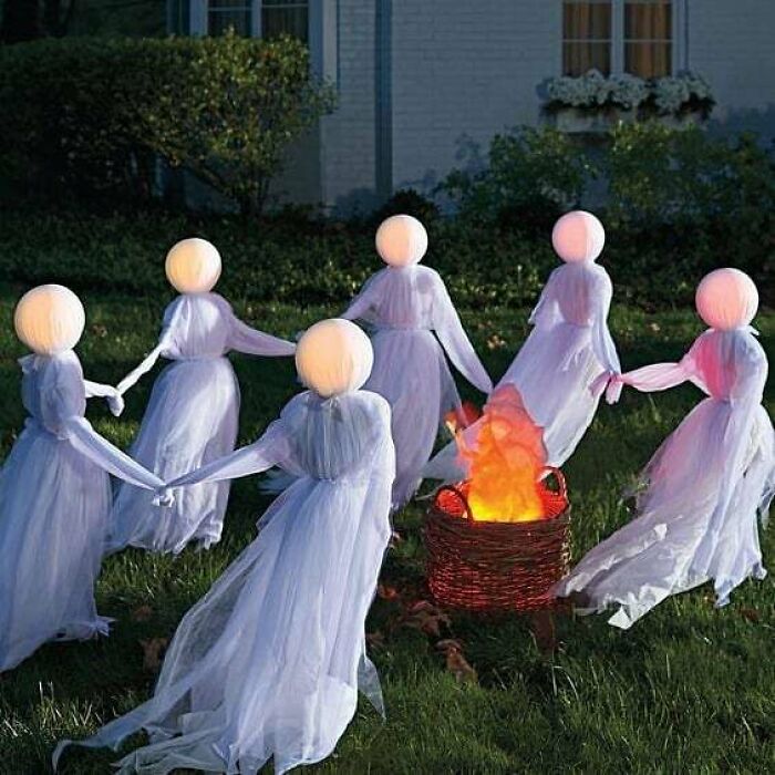 Outdoor Halloween decorations featuring glowing ghost figures holding hands around a flickering fire basket on a lawn at night