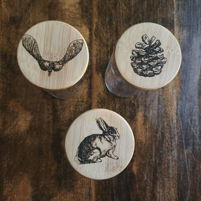 Three wooden lids with wood-burned art featuring a rabbit, pinecone, and seed pod by Canadian artist Kat Slota.