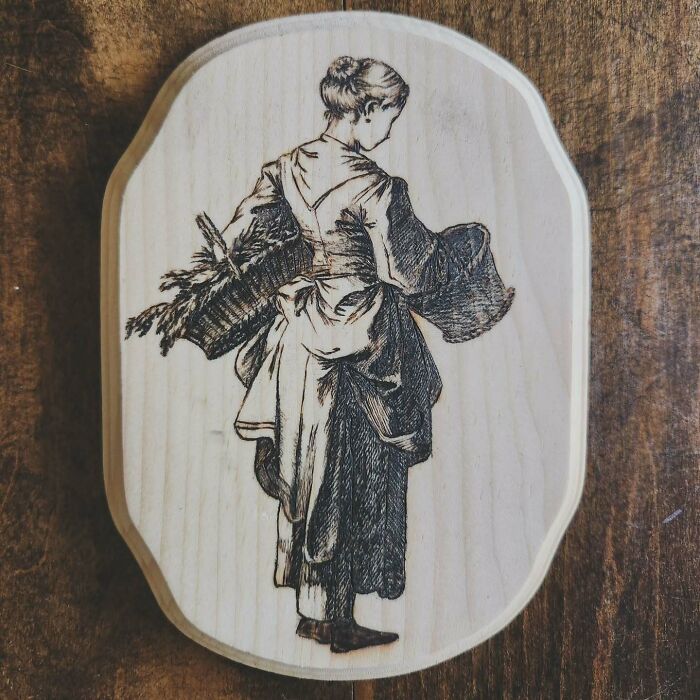 Wood-burned artwork of a woman carrying a basket, created by Canadian artist Kat Slota using fire on a wooden plaque.