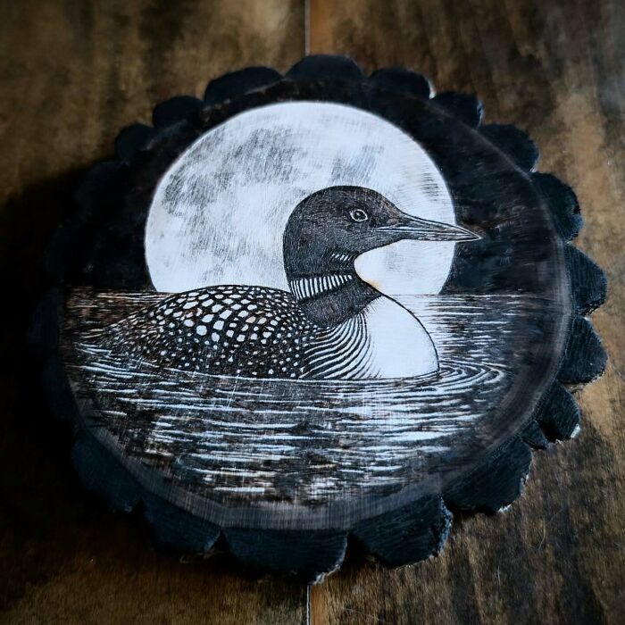 Wood-burned artwork of a loon swimming at night with a full moon, created by Canadian artist Kat Slota using fire.