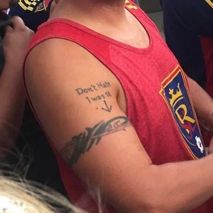 Man wearing red tank top showing arm tattoo with text apologizing for poor decision at 18, example of awful tattoos.