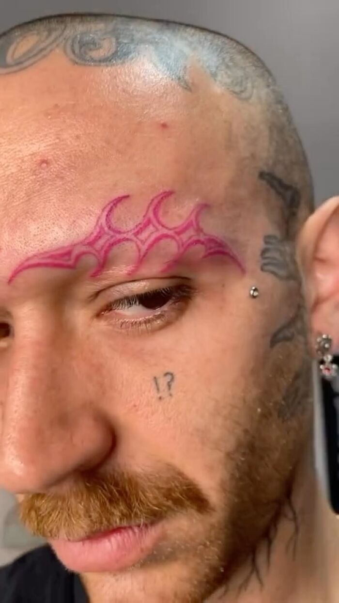 Close-up of a man with awful face tattoos including a pink tribal design above the eyebrow and a small punctuation mark on the cheek.