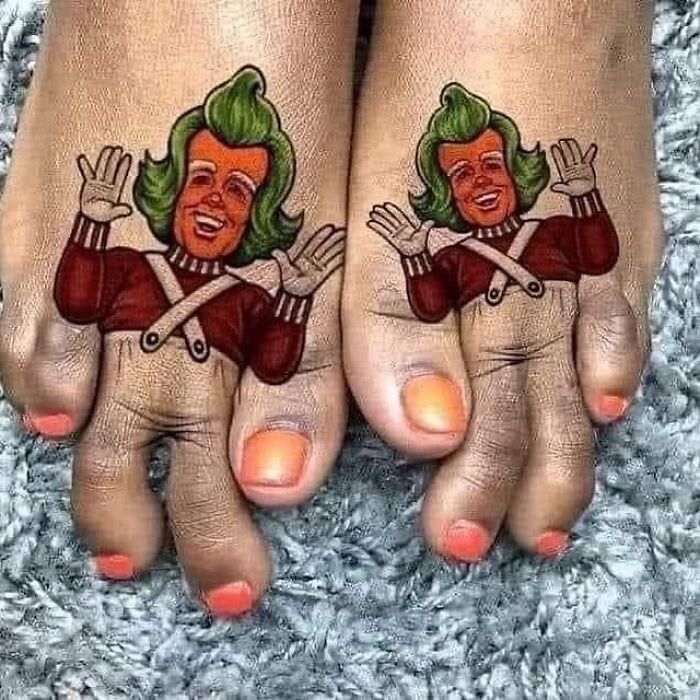 Matching colorful awful tattoos of a cartoon character on toes with painted orange nails on a gray carpet background.