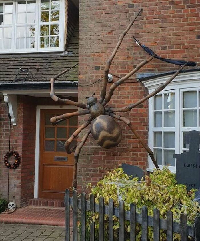 Large outdoor Halloween decoration of a giant spider attached to brick house wall near Halloween-themed yard display.