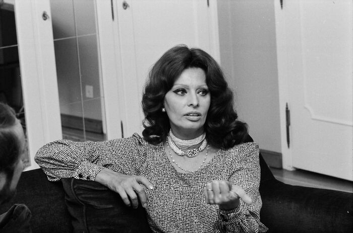 Sophia Loren seated on a couch, wearing a patterned blouse and layered necklaces, engaged in conversation indoors.