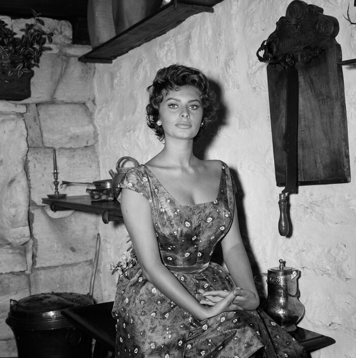 Sophia Loren sitting in a vintage room, showcasing Italian beauty and classic Hollywood style in a floral dress.