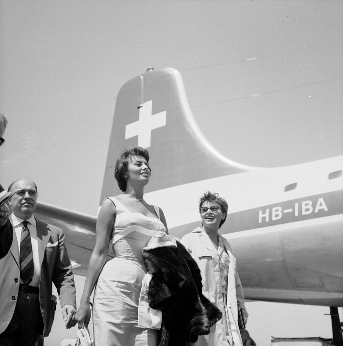 Sophia Loren standing by a Swiss airplane tail, dressed elegantly, reflecting classic Italian beauty and Hollywood legend charm.