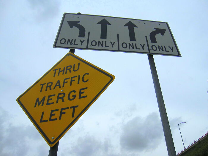 Confusing road signs showing multiple directional arrows and a merge left warning under cloudy sky background.
