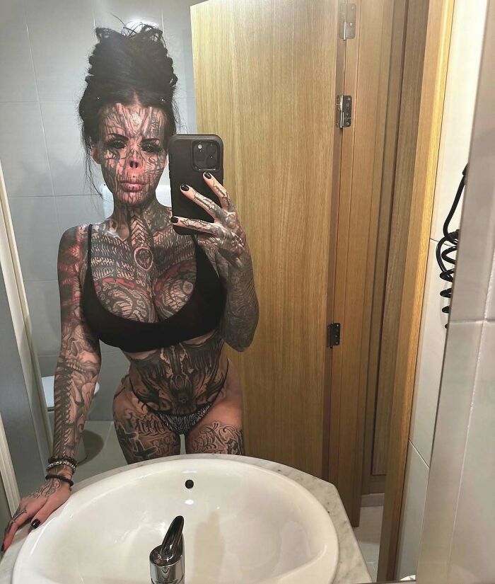 Tattooed woman with extensive body art takes a mirror selfie wearing black underwear in a bathroom setting.