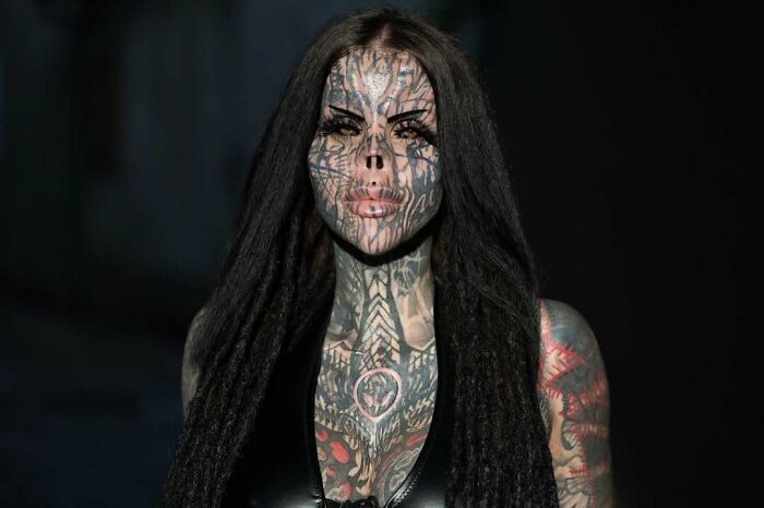 Tattooed woman with extensive facial and body tattoos posing against a dark background, showcasing unique body art.