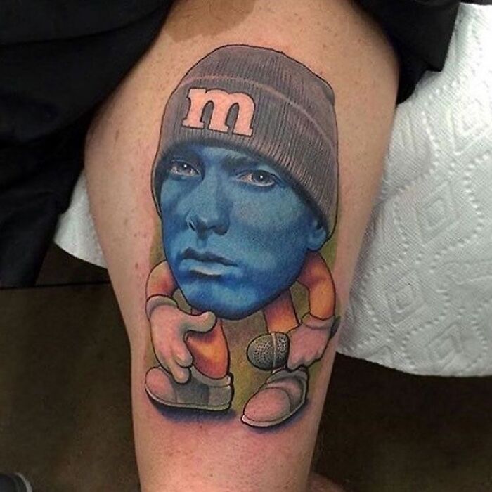 Tattoo of a blue-faced character wearing an M&M beanie, with cartoon arms and legs holding a microphone on a thigh.