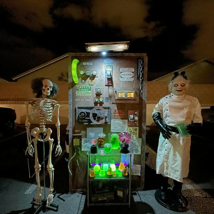 Outdoor Halloween decorations featuring a glowing mad scientist display with skeleton and spooky lab equipment at night