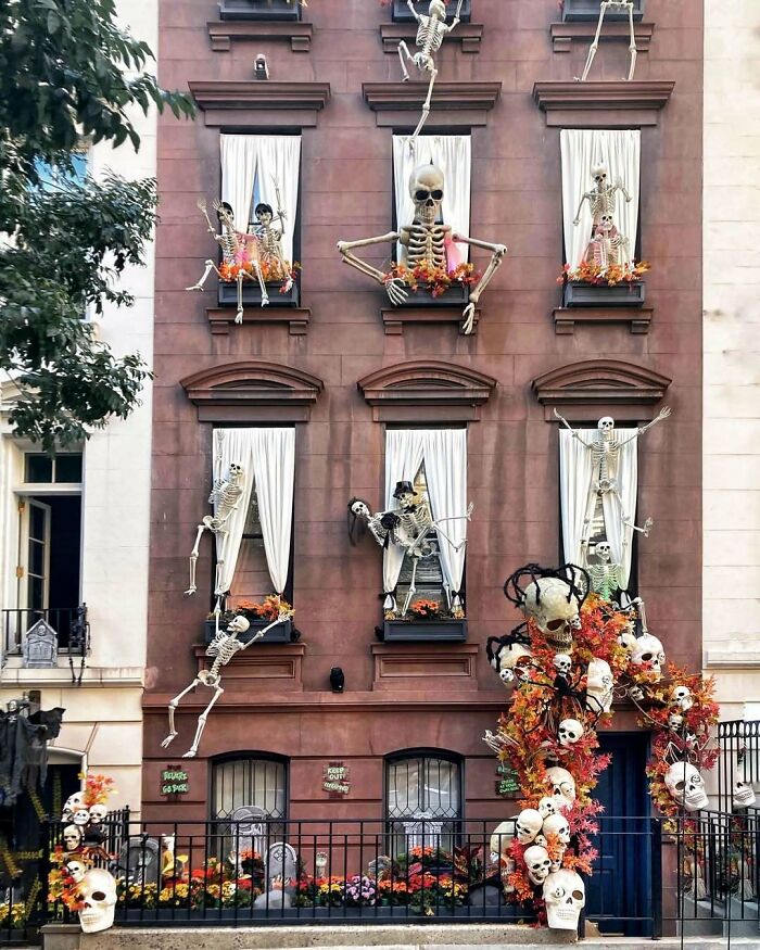 Large outdoor Halloween decorations featuring multiple skeletons displaying spooky antics on a building facade with fall foliage.