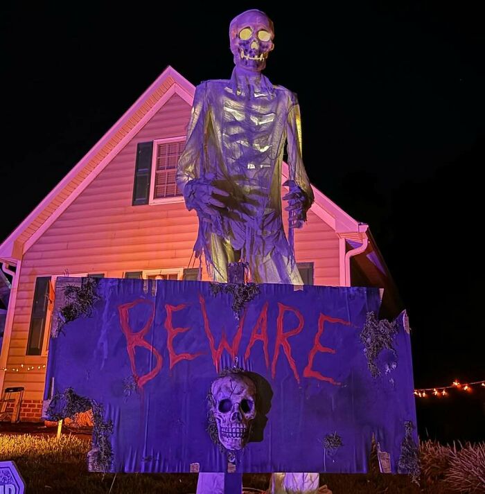 Giant skeleton with glowing eyes and beware sign in front of house as outdoor Halloween decoration at night.