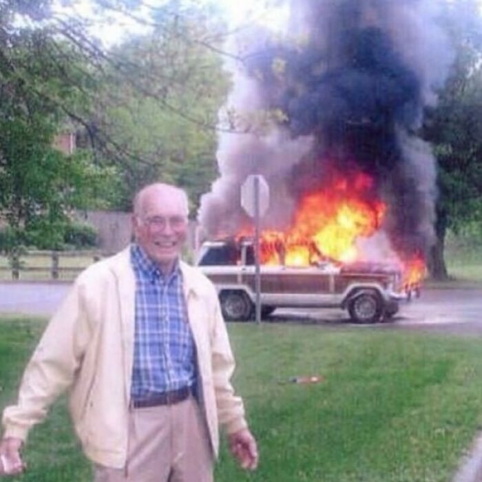 Elderly man smiling in front of a burning car with flames and thick black smoke, a scene that might need a priest or exorcist.