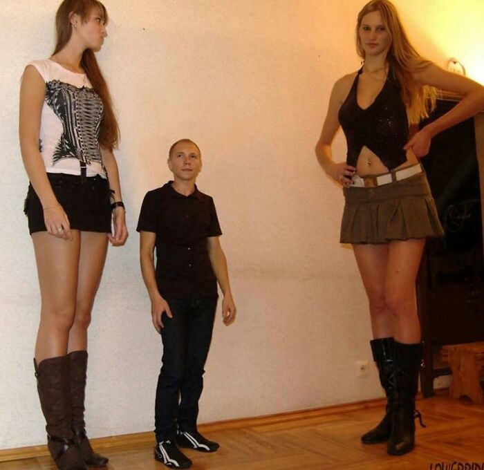 Two tall women and a much shorter man standing indoors with wood flooring, an unusual height difference capturing attention.