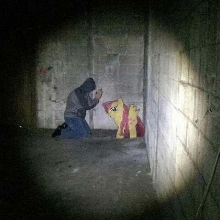 Person kneeling in a dark, concrete room facing a cartoon pony, an eerie scene that might need a priest or an exorcist.
